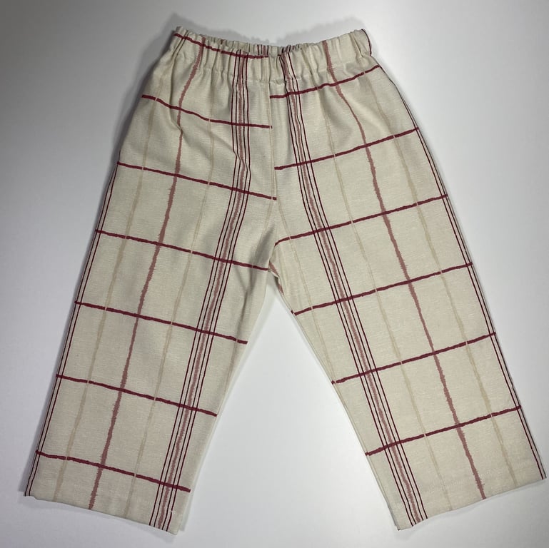 Bukser i cremefarvet stof med elastisk talje og ternet mønster i røde og beige nuancer, lagt fladt på en hvid baggrund.