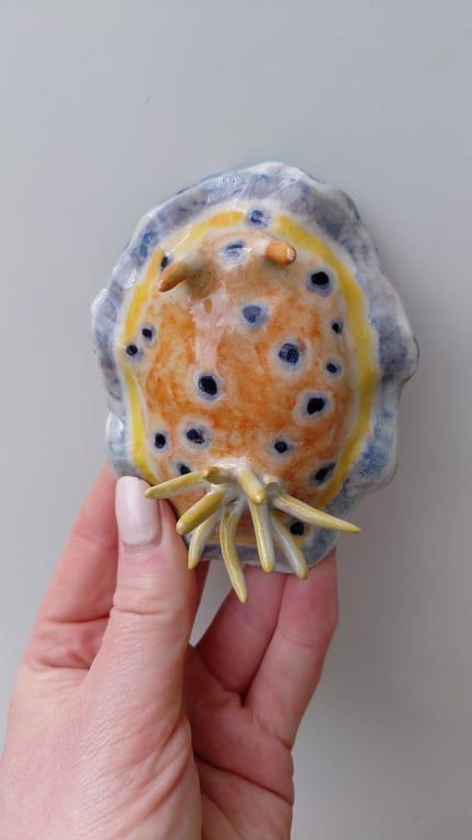 Hånd, der holder en håndlavet keramisk figur af en orange og blå pletskørbug med gule tentakler.