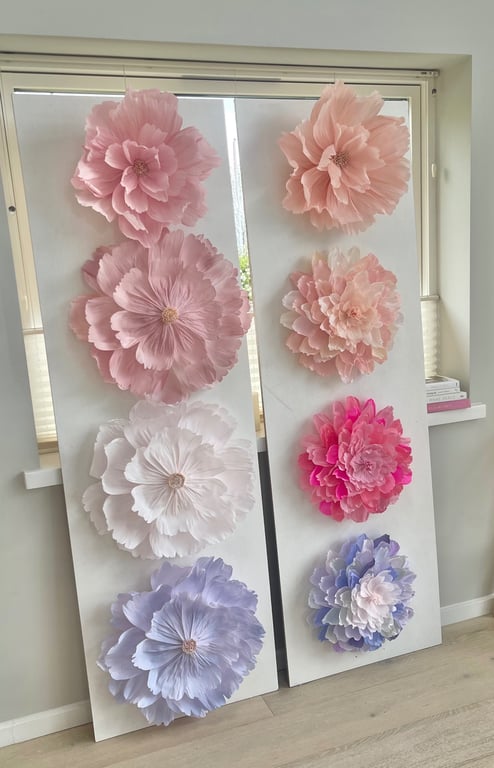To store two white display boards each decorated with six large, layered papirblomster i forskellige farver, herunder lyserød, hvid, lilla og pink, placeret foran et vindue i et lyst rum.