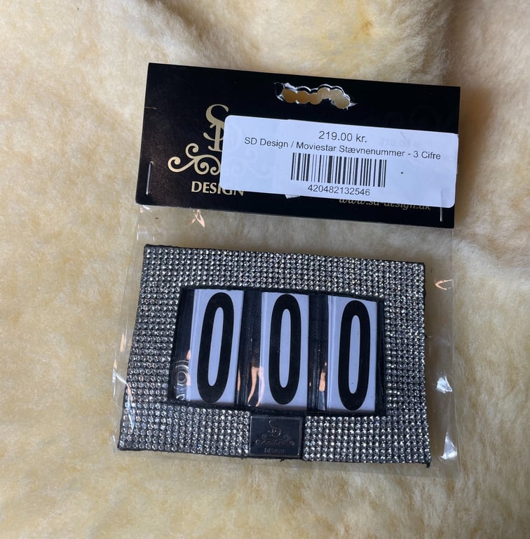 Pakke med glitrende, diamantbesat nummerplade med tallet "000" fra SD Design, pris 219 kr., på blød, cremefarvet baggrund.