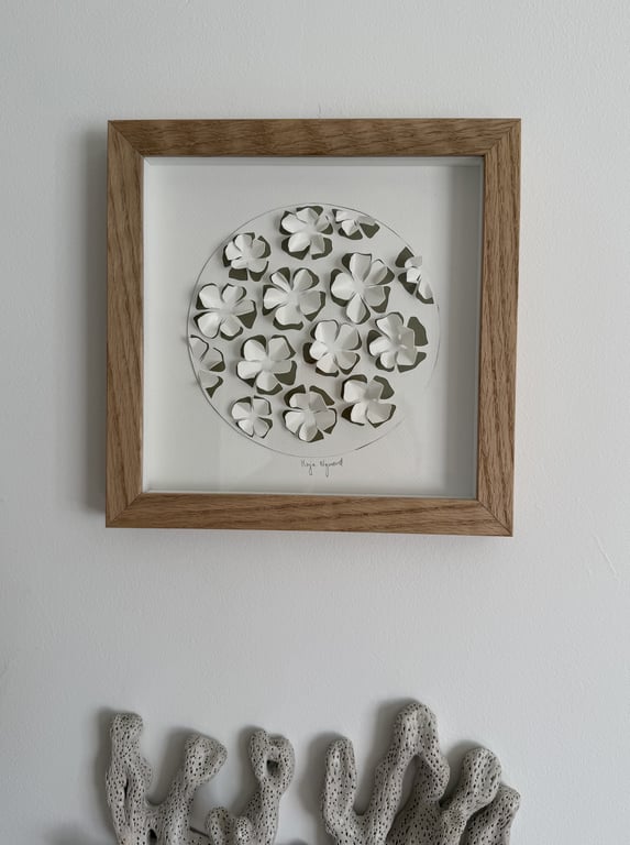 Billedramme med udskårne hvide papirblomster i en cirkel, hængende på en hvid væg over en struktur med porøst mønster.