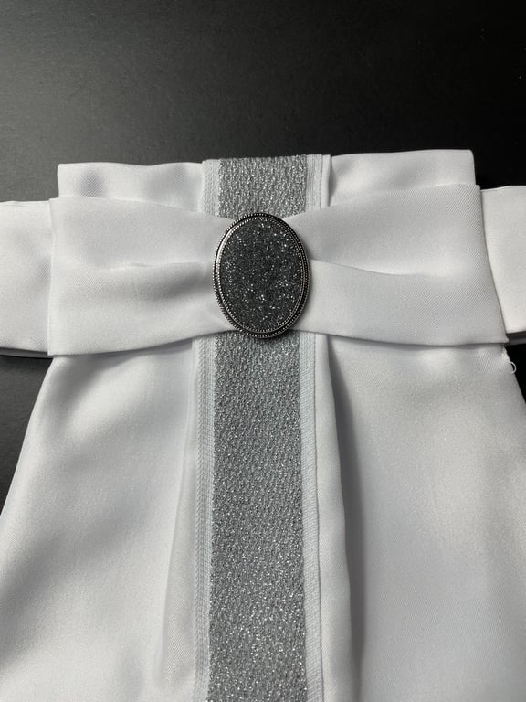 Hvid kjole med sølvglitrende bånd og stor oval sølvbroche ved taljen.