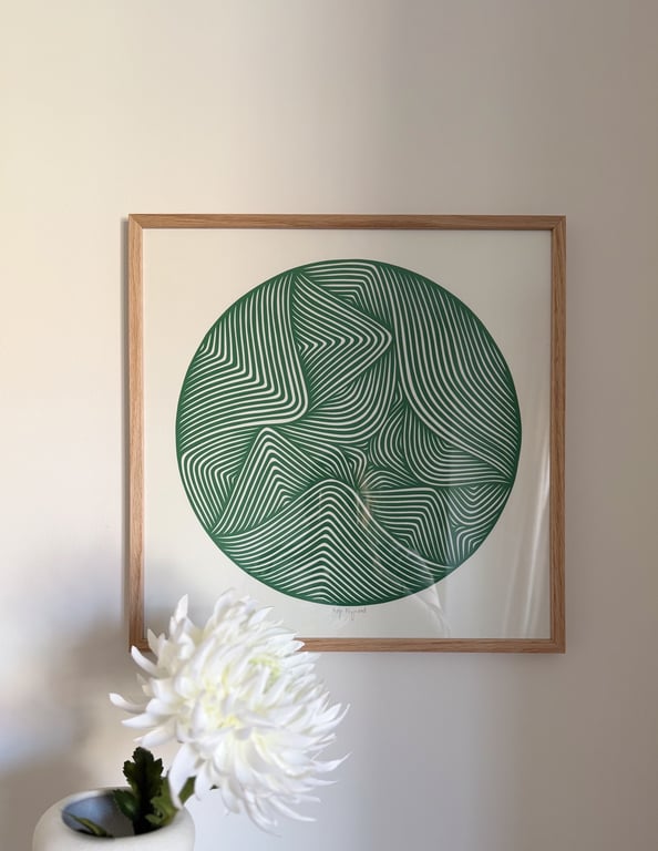 Indrammet abstrakt kunst med grønne, bølgede linjer i en cirkel på hvid baggrund, hængende på en beige væg med en hvid blomst i forgrunden.