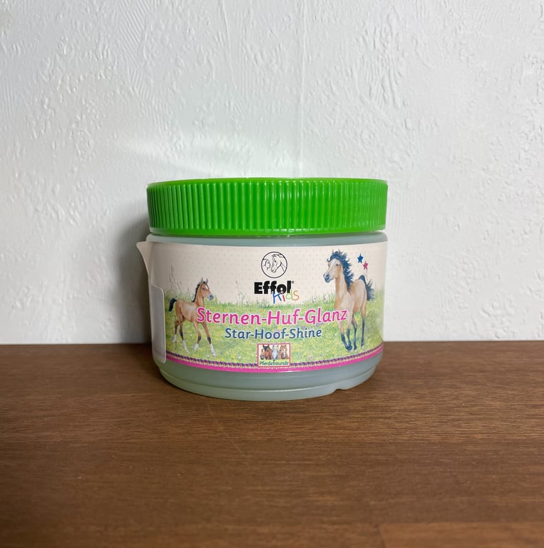 Plastbeholder med grønt låg mærket "Effol Kids Sternen-Huf-Glanz Star-Hoof-Shine" med illustrationer af heste, stående på en træoverflade foran en hvid væg.