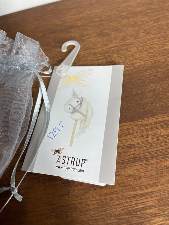 Et produktmærkat med en illustration af en hvid hobbyhest med mane på en pind, mærket har teksten "ASTRUP" og en pris på 129 kr., ligger på et træbord ved siden af en gennemsigtig organzapose med en stålgrå snor og en lille plastikbøjle.