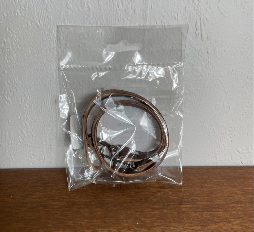 Plastpose med to brune læderremme med metalspænder, placeret på en træflade foran en hvid væg.