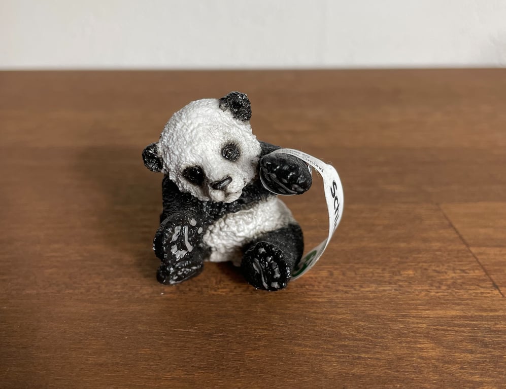 Lille sort-hvid pandafigur i plastik med et hvidt mærke, placeret på et brunt træbord.