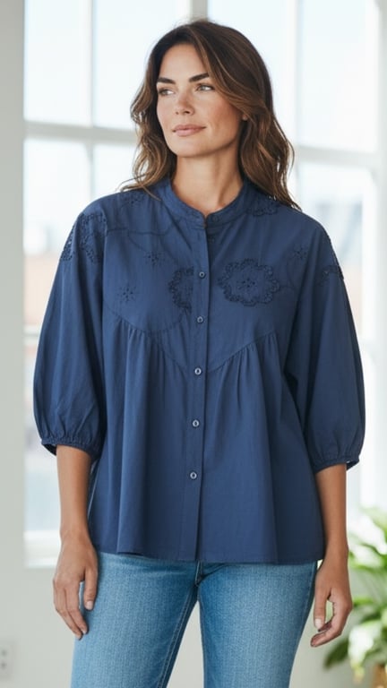 Kvinde iført mørkeblå bluse med broderi og lyseblå jeans, stående foran lyst vindue.