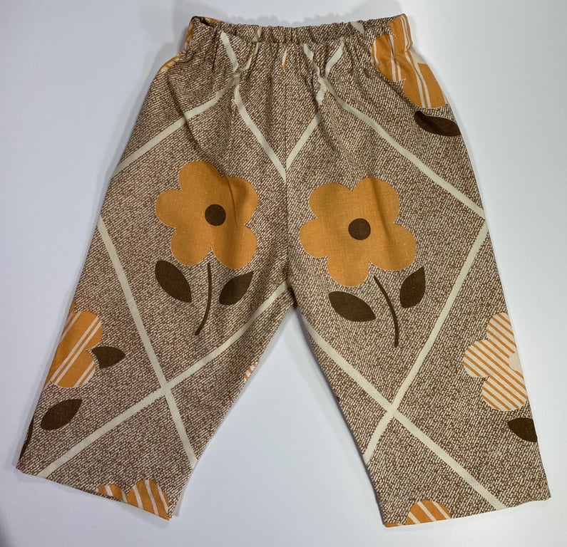 Børnebukser med elastik i taljen og mønster af store orange blomster og diagonale striber på brun baggrund.