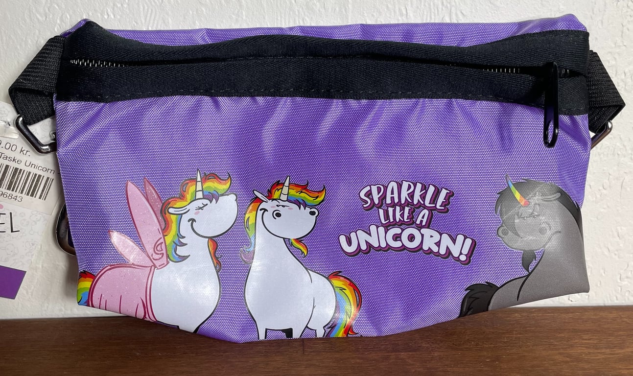 Lilla taske med sort lynlås, pyntet med tegninger af hvide og grå enhjørninger med regnbuefarvede manker og teksten "Sparkle like a unicorn!"