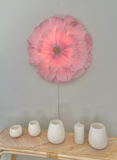 Lysende væglampe formet som en stor pink blomst, hængende over et bambusbord med fem hvide lys og fyrfadslysholdere. (1)