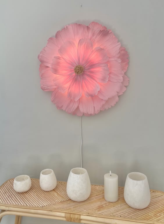 Lysende væglampe formet som en stor pink blomst, hængende over et bambusbord med fem hvide lys og fyrfadslysholdere.