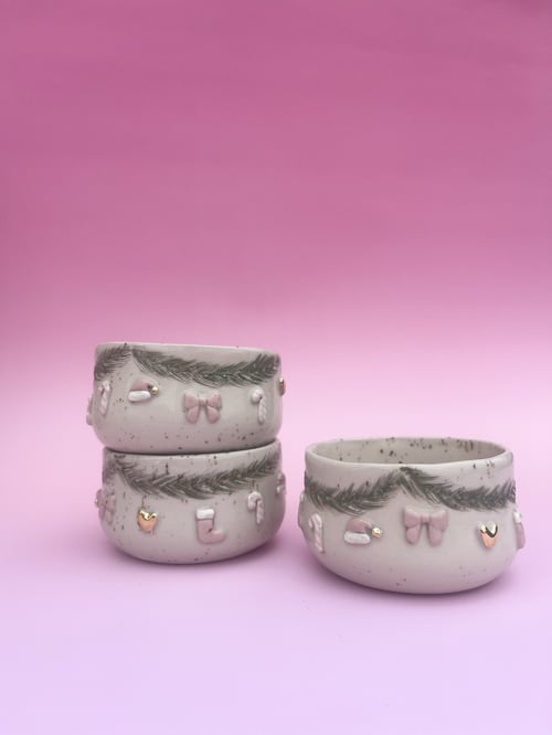 Tre keramikskåle med juledekor på, herunder guirlander, sløjfer, hjerter og julestrømper, mod en pink baggrund. (1)