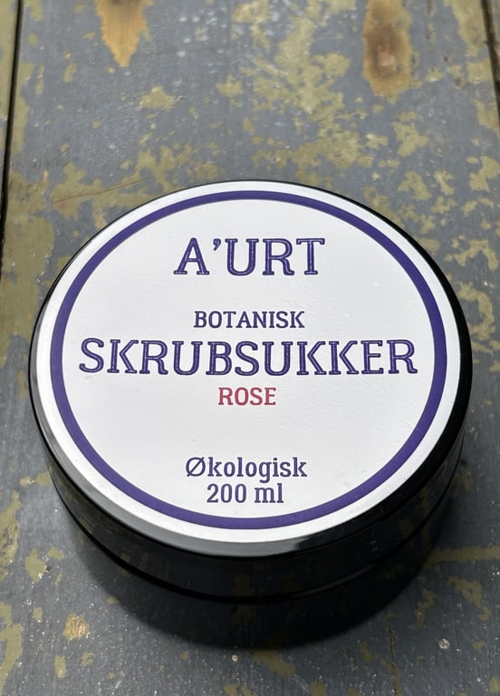 Dåse med A'URT botanisk skrubsukker med rose, økologisk, 200 ml, placeret på en slidt, blågrå træflade.