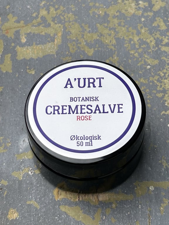 Sort krukke med hvidt mærkat, der viser navnet "A'URT Botanisk Cremesalve Rose" og angiver, at produktet er økologisk og indeholder 50 ml.