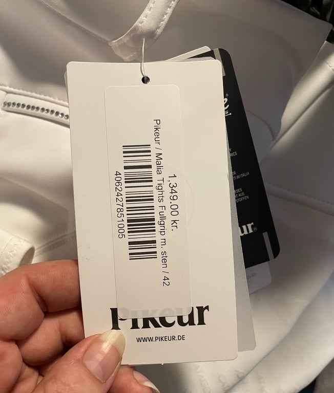 Et hånd, der holder en pris- og produktmærkat for Pikeur Malia Tights Fullgrip i stenfarve, størrelse 42, med prisen 1.349,00 kr. mærket er fastgjort til et lyst stykke tøj.