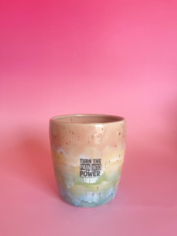 Keramikkrus med farvet glasur i nuancer af beige, gul, grøn og blå foran en pink baggrund med teksten "TURN THE PAIN INTO POWER".