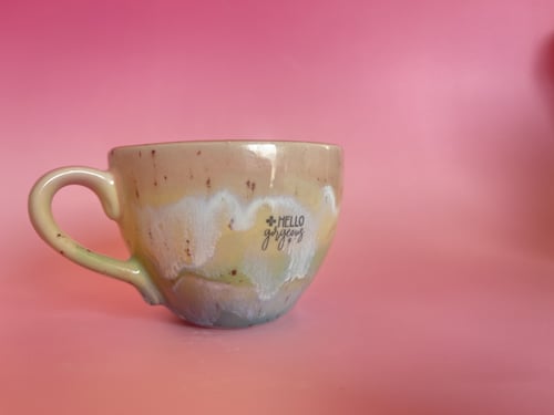 En beige keramisk kop med glasur i forskellige nuancer og teksten "HELLO gorgeous" mod en pink baggrund. (1)