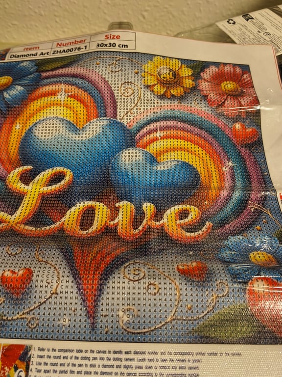Billede af et diamantmaleri med to blå hjerter og regnbuer i baggrunden, med teksten "Love" i forgrunden samt farverige blomster omkring.