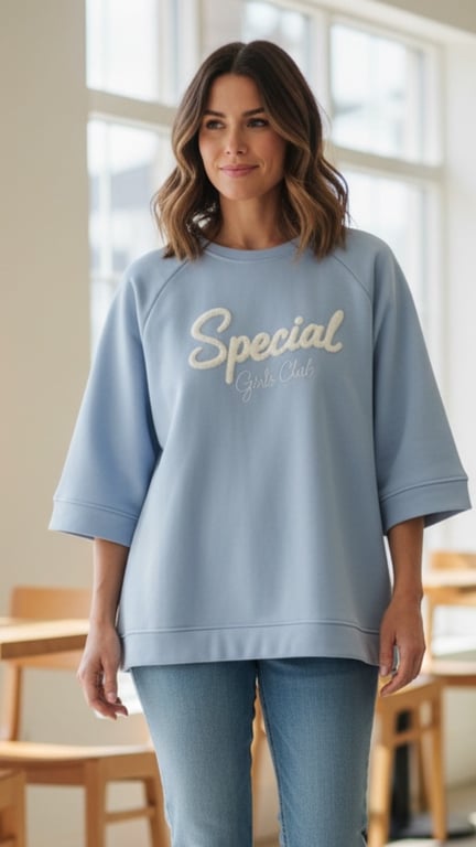 Kvinde iført lyseblå sweatshirt med teksten "Special Girls Club" og lyseblå jeans, stående i lyst rum med vinduer og træmøbler.