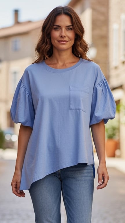 Kvinde iført lyseblå bluse med pufærmer og asymmetrisk kant samt blå jeans, gående udendørs i bymiljø.