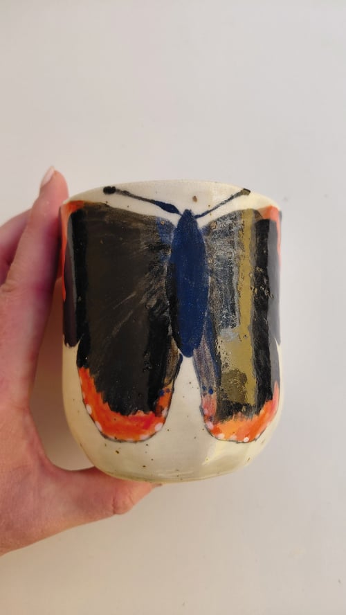 Hånd der holder en keramikkrus med malet motiv af en sommerfugl med sorte, blå og orange vinger. (1)
