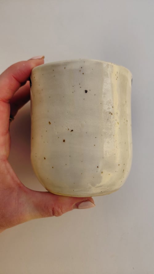 Hånd, der holder en hvid keramikkop med små mørke prikker mod en neutral baggrund. (4)