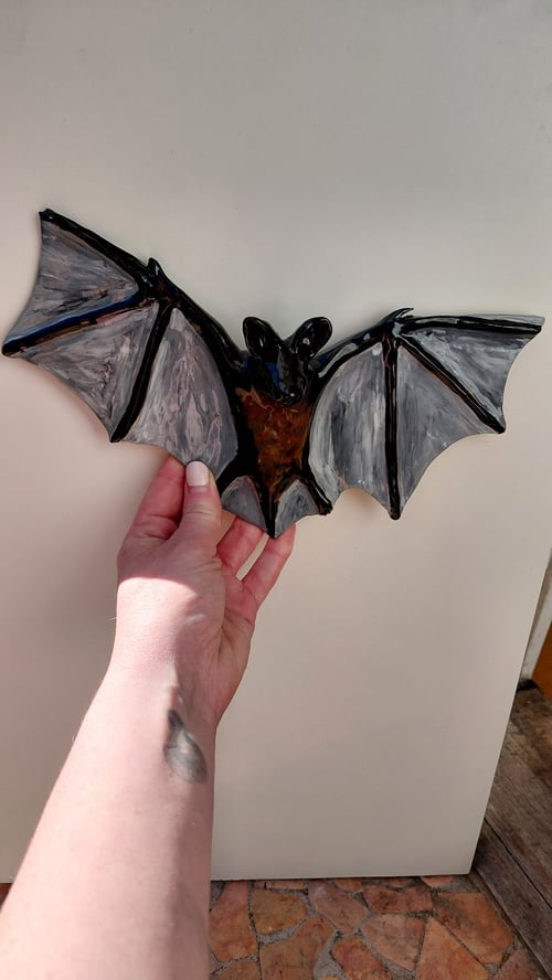 Hånd, der holder en sort og grå flagermusfigur mod en hvid væg. (1)