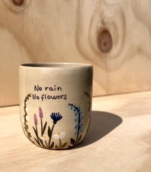 Beige keramikkrus med håndmalede blomster og teksten "No rain No flowers" stående på lyst træbord med lyst træbaggrund. (1)