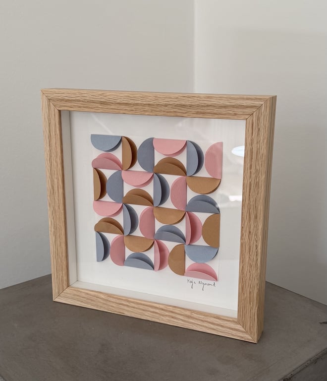 Indrammet abstrakt kunstværk bestående af farvede halvcirkler i blå, rosa og brun, arrangeret i et geometrisk mønster.