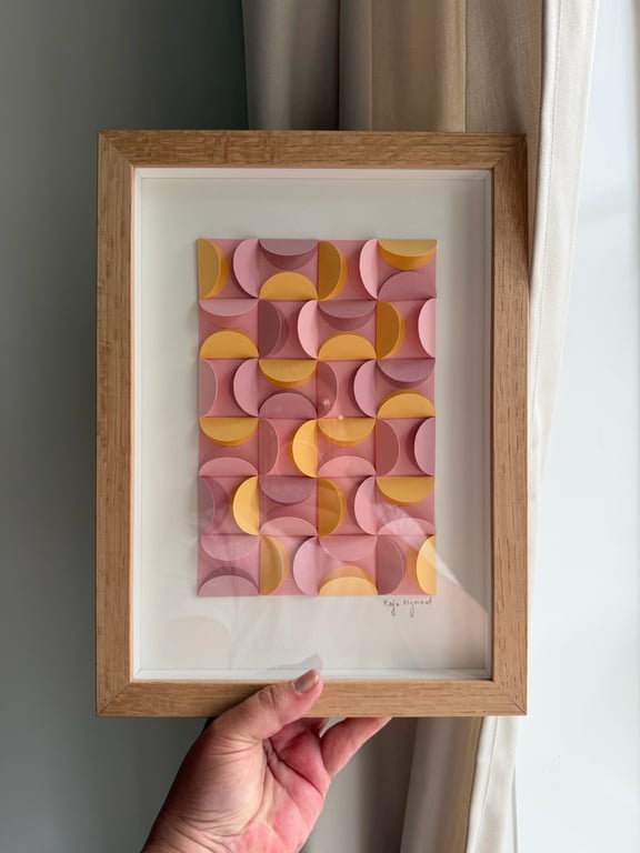 Hånd, der holder en indrammet abstrakt papirkunst med geometriske former i rosa og gule nuancer.