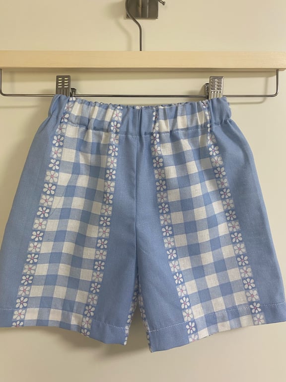 Lyseblå og hvide ternede shorts med blomstermønster, hængende på en bøjle mod en neutral baggrund.