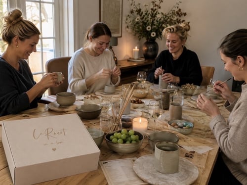 Fire kvinder sidder rundt om et bord og laver keramik sammen, omgivet af snacks, levende lys og vin. (1)