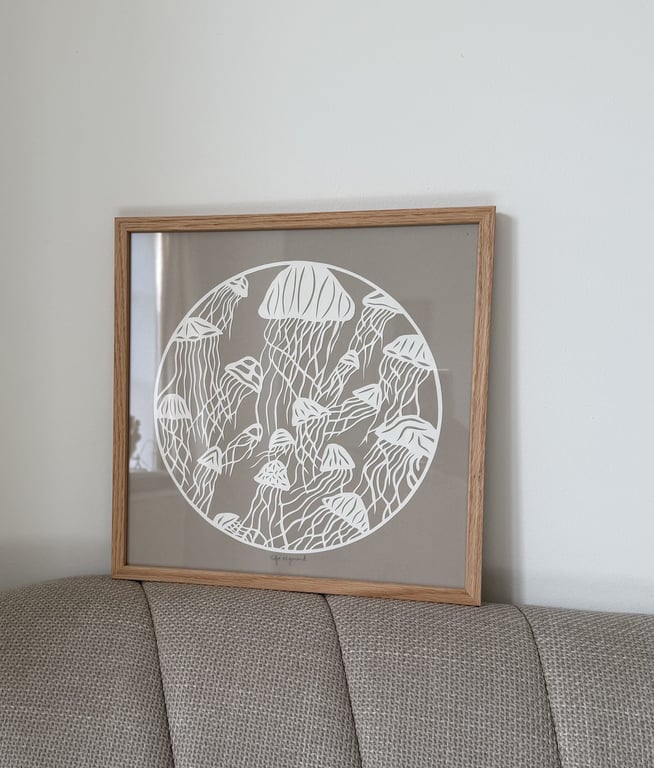 Billedramme med hvid illustration af vandmænd i en cirkel på beige baggrund, hængende over en grå stofsofa mod en hvid væg.