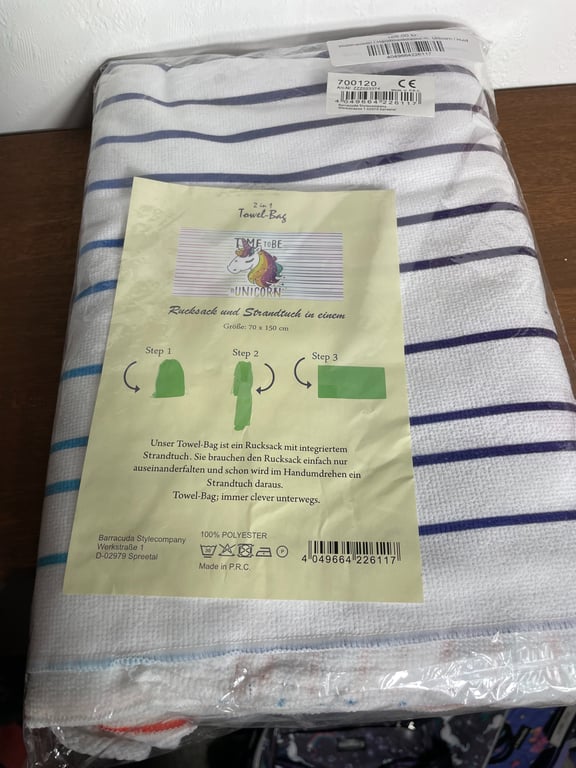 Foldet hvidt håndklæde med blå striber i emballage med et beige informationsark på, der beskriver en 2-i-1 towel-bag med illustration af en enhjørning og instruktioner på tysk.