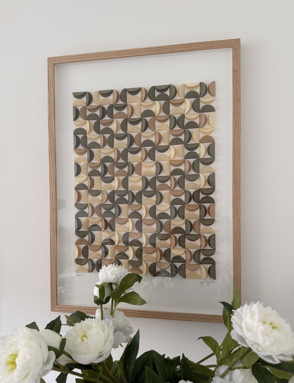 Abstrakt vægkunst bestående af mønstrede, buede firkanter i beige, brune og grønne nuancer, indrammet i lyst træ, med hvide blomster i forgrunden.