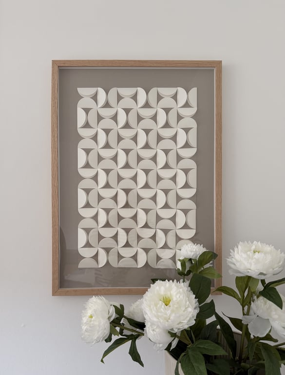 Minimalistisk vægkunst bestående af geometriske hvide former i et rektangulært lyst træindrammet billede, med hvide blomster i forgrunden mod en lys væg.