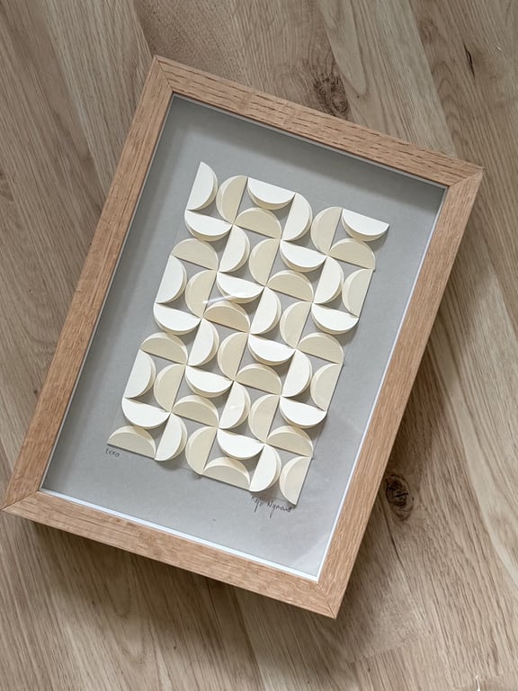 Billedramme med geometrisk papirkunst bestående af buede, foldede hvide og beige former arrangeret i et mønster på grå baggrund, placeret på trægulv.