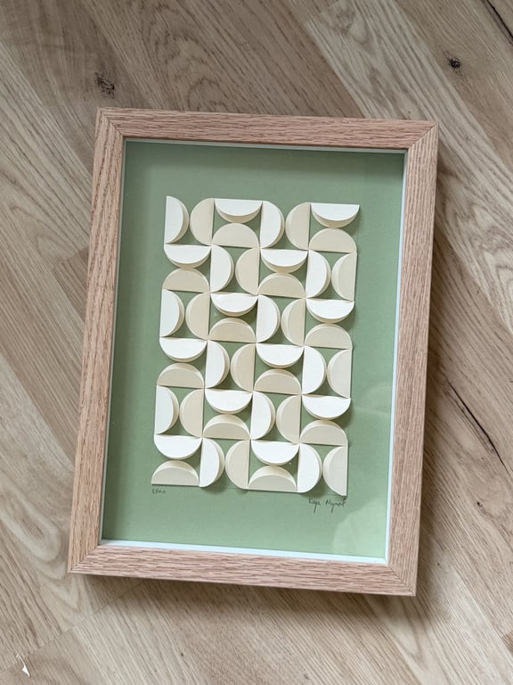 Indrammet abstrakt papirkunst med geometriske, halvcirkelformede mønstre i beige og hvidt på grøn baggrund, placeret på trægulv.