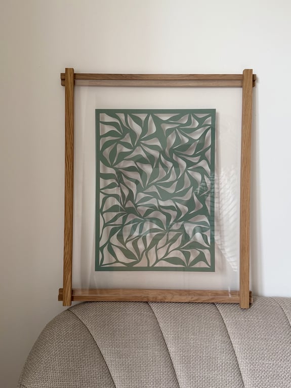 Billedramme med trækant og et kunstværk bestående af grønne, abstrakte blade på transparent baggrund, hængende på en hvid væg over en beige sofa.