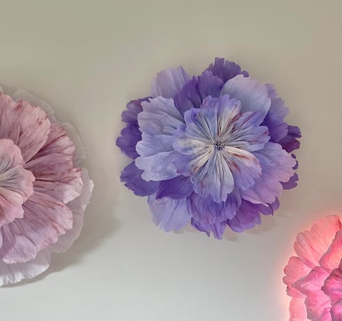 Tre store, kunstige blomster i lyserøde og lilla nuancer fastgjort på en lys væg. (1)