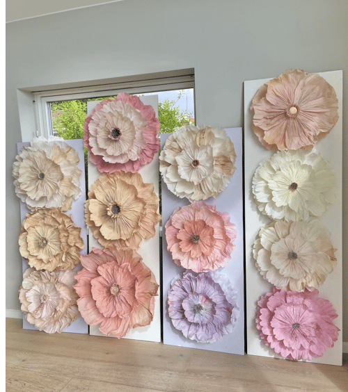 Store blomster lavet af papir i bløde farver som lyserød, beige, hvid og lavendel, monteret på lodrette hvide plader, stående på et trægulv foran et vindue. (1)