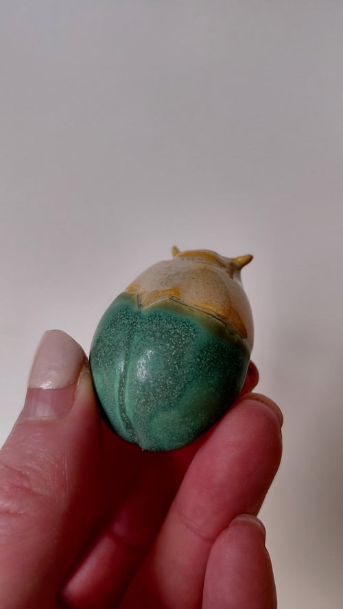 Hånd der holder en lille, glaseret keramisk figur formet som en grøn og brun hasselnød. (1)