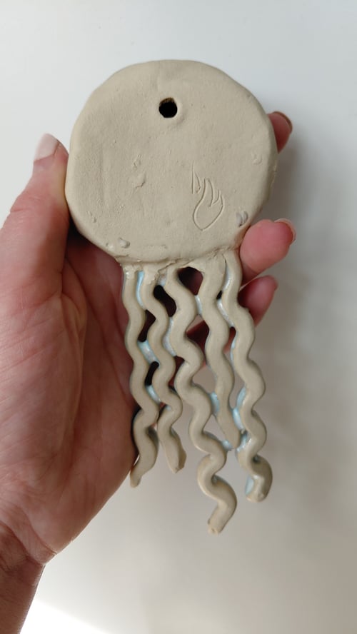 Hånd holder en keramisk figur, der ligner en blæksprutte med otte bølgede ben, beige farve og et hul i toppen. (7)