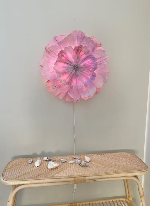 Pink blomstformet væglampe tændt over et lyst bambusbord med små sten og skaller på. (1)
