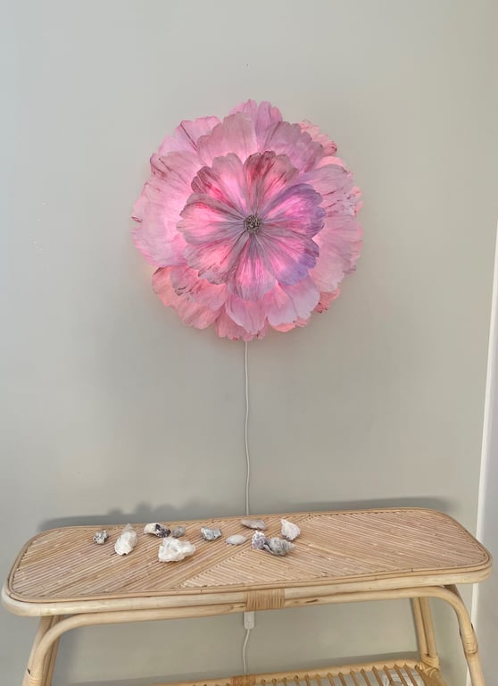 Pink blomstformet væglampe tændt over et lyst bambusbord med små sten og skaller på.