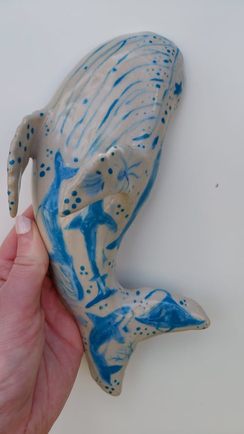 Hånd, der holder en hvalformet keramikfigur med blå malede havdyr og mønstre på beige baggrund. (1)