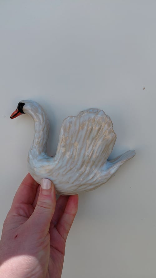 Hånd, der holder en keramisk figur af en svane med spredte vinger mod en lys baggrund. (1)