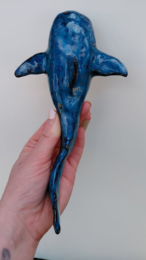 Hånd holder en keramisk blå hajfigur set oppefra mod en lys baggrund. (1)