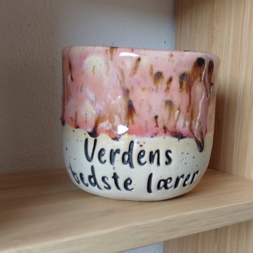 Keramikkop med rosa glasur og teksten "Verdens bedste lærer" placeret på en træhylde. (1)